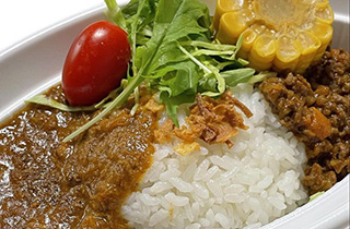 写真：ご飯を真ん中にして両側にチキンカレーとキーマカレーが盛られている。ご飯とルーの上にはミニトマトと緑の野菜、とうもろこしがのっている